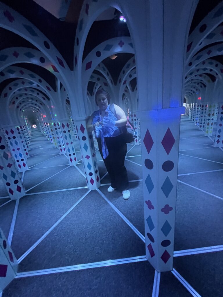 אדם עומד במבוך מראות עם קשתות לבנות המעוטרות בדוגמאות צבעוניות של יהלומים וקלפים, היוצרות השתקפויות מרובות ואשליה אופטית של מעברים אינסופיים.