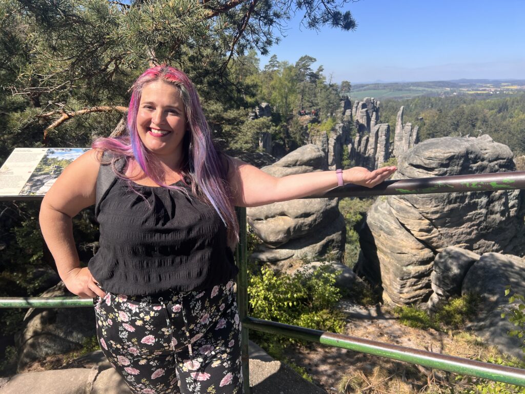 אישה מחייכת עם שיער ורוד וסגול עומדת ליד מעקה מתכת המשקיף על צוקים סלעיים ונוף מיוער ביום שטוף שמש. היא לובשת חולצה שחורה ומכנסיים פרחוניים.