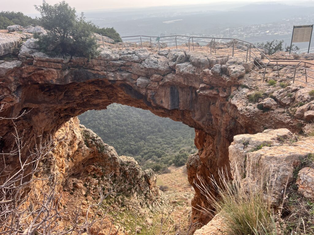קשת מאבן טבעית משתרעת על צוק מחוספס מעל עמק ירוק, עם מעקה בטיחות מתכתי לאורך החלק העליון ועצים ושיחים הצומחים בסמוך תחת שמיים מעורפלים.