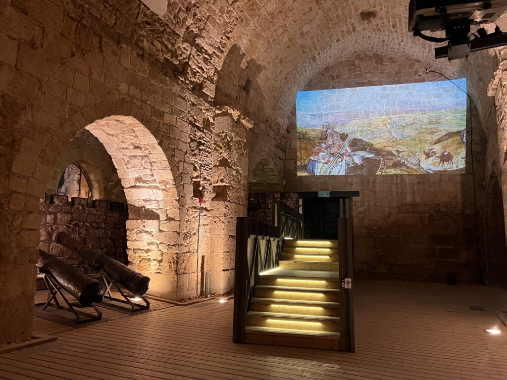 חדר עם קירות אבן ותקרות מקושתות, ובו שני תותחים עתיקים משמאל ומדרגות מוארות במרכז. תמונה היסטורית צבעונית מוקרנת על הקיר מעל המדרגות.