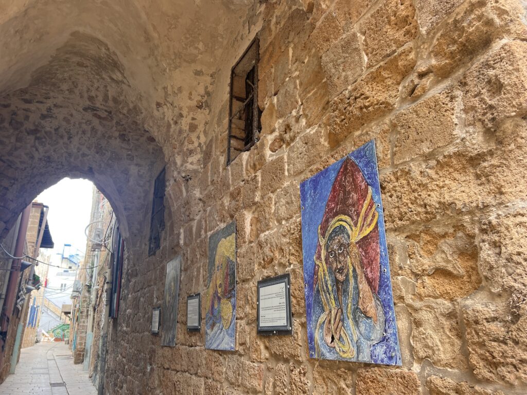 סמטה צרה מאבן עם תקרה מקושתת, מעוטרת בציורים צבעוניים ולוחות המוצבים על קיר הלבנים העתיק. אור השמש מסתנן מבעד לקשת ומאיר את האבנים המחוספסות ואת יצירות האמנות.