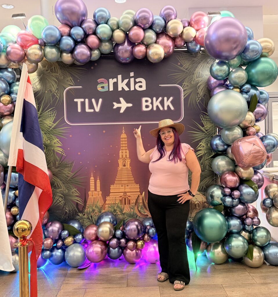 אישה לבושה בחולצה ורודה וכובע עומדת מחייכת מול קשת בלונים צבעונית עם שלט שעליו כתוב arkia TLV → BKK, עם איורים של בנגקוק ודגל תאילנד סמוך.