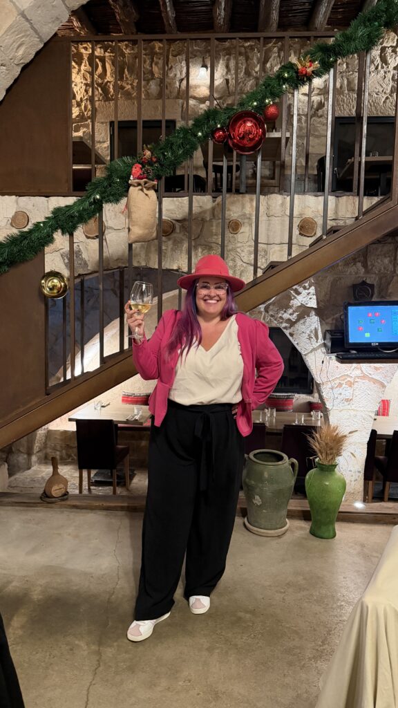 אדם מחייך עם שיער סגול ארוך, חובש כובע ורוד ולובש ז'קט, חולצה לבנה ומכנסיים שחורים, אוחז בכוס יין. הוא עומד בתוך הבית ליד גרם מדרגות מעוטר בזר פרחים ובקישוטים.