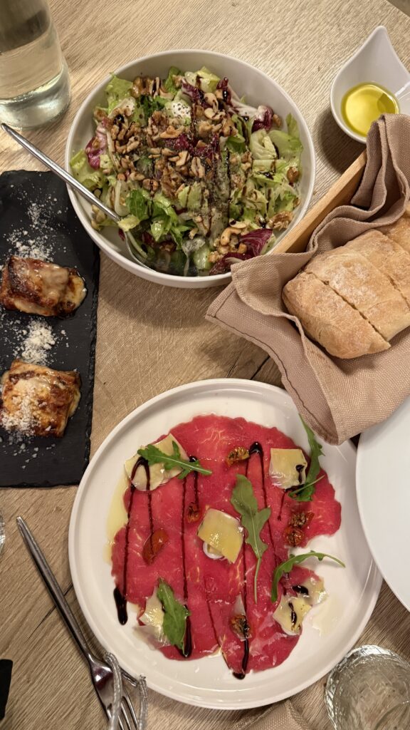 שולחן ערוך עם סלט ירוק טרי, לחם עם שמן זית, שני מאפים קטנים עם גבינה, וצלחת קרפצ'יו בקר מעוטר ברוקט, פרוסות גבינה, עגבניות מיובשות וזיגוג בלסמי.