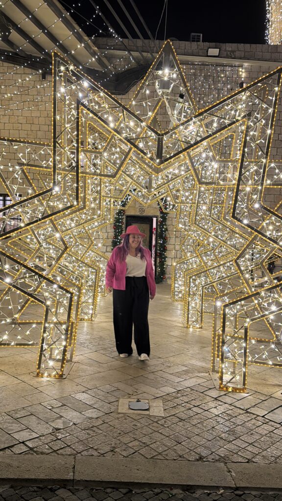 אדם לבוש ז'קט ורוד ומכנסיים שחורים עומד מחייך מתחת למנהרה גדולה בצורת כוכב, מקושטת באורות חג לבנים וצהובים בוהקים, על רקע בניין אבן בלילה.