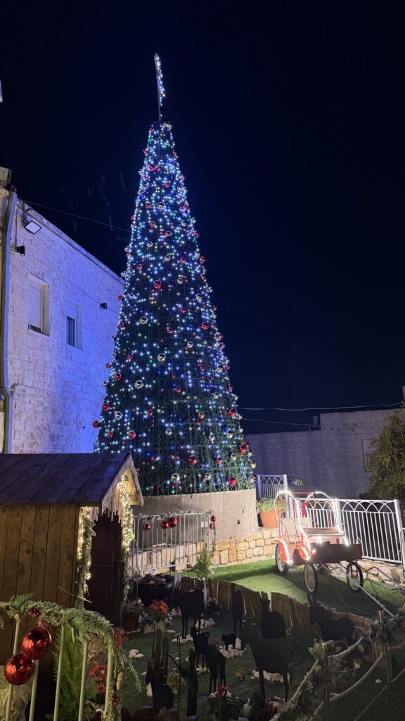 עץ חג מולד גבוה ומואר, מקושט באורות צבעוניים ובקישוטים, ניצב בלילה בחוץ לצד בניינים. מלפנים נראים סצנת לידת ישו קטנה ומכונית צעצוע אדומה, מוקפות בקישוטים חגיגיים.