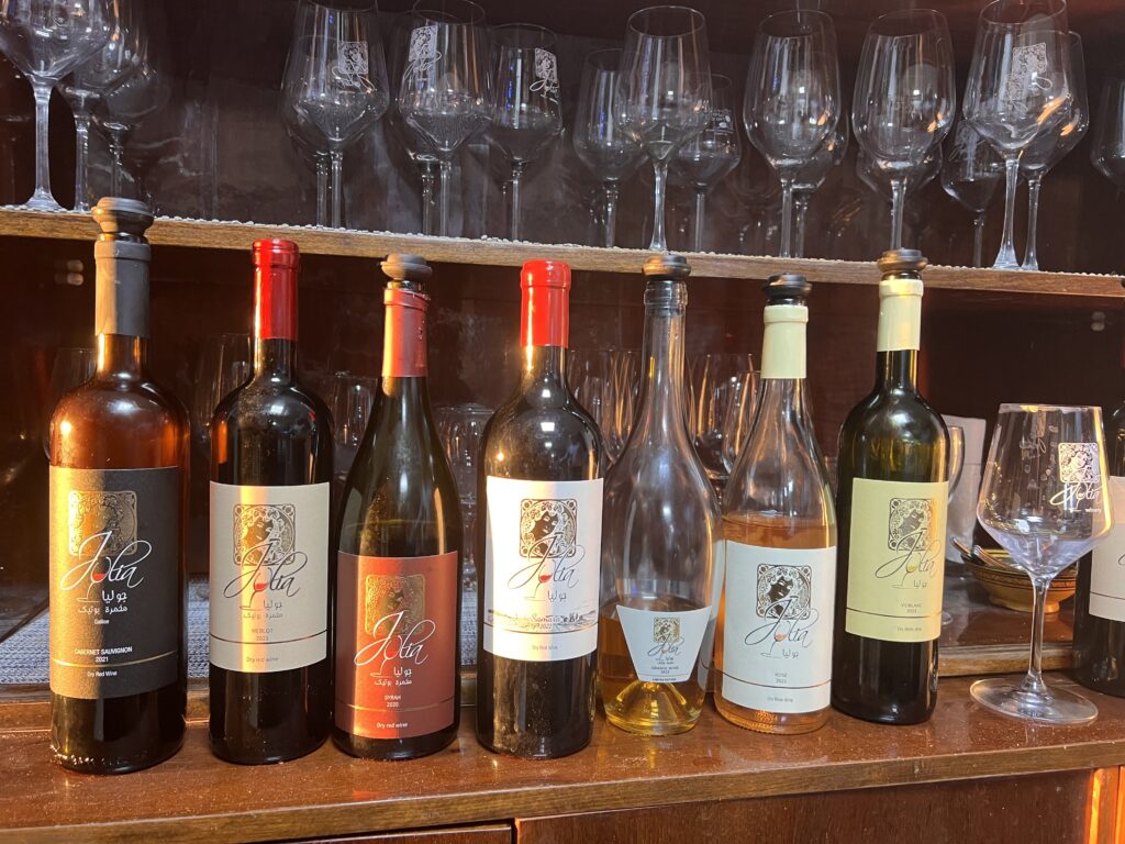 שבעה בקבוקי יין עם תוויות בצבעים שונים מסודרים על משטח עץ, וכוסות יין וגביעים ריקים מסודרים על מדפים מאחוריהם. תאורה חמימה מדגישה את הבקבוקים והכוסות.