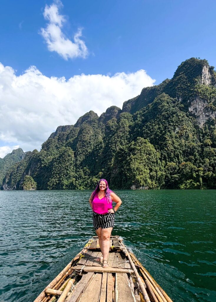 אישה עם שיער סגול עומדת על רפסודה מעץ הצפה על אגם, מוקפת בהרים גבוהים וירוקים תחת שמיים כחולים עם עננים פזורים.