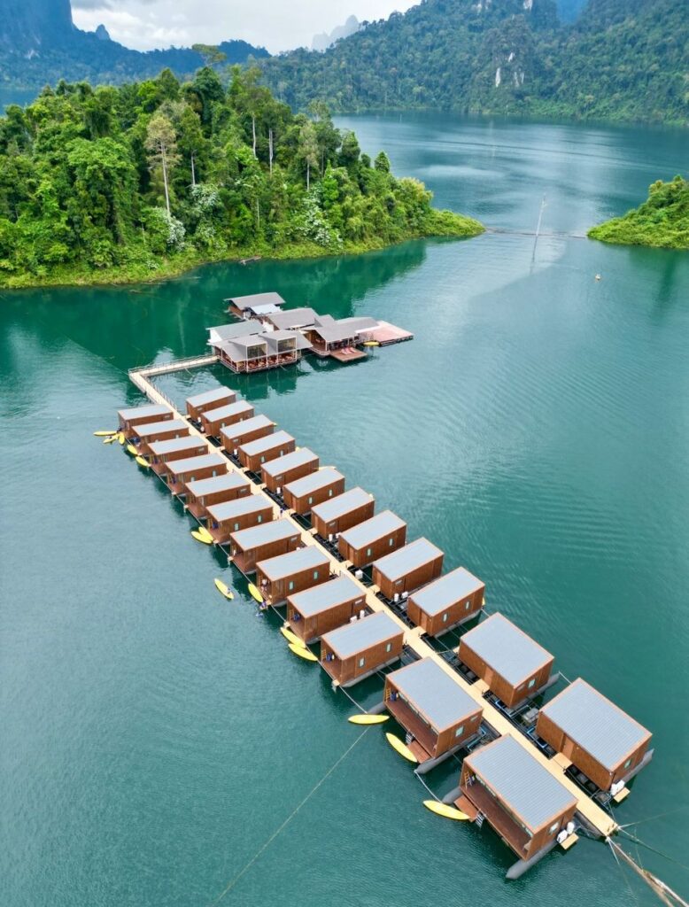 בונגלוס צפים מסודרים בשורות על אגם שליו, מוקפים ביער ירוק ושופע ובהרים, עם קיאקים עוגנים בכל יחידה וקבוצת מבנים מרכזיים בקרבת מקום.