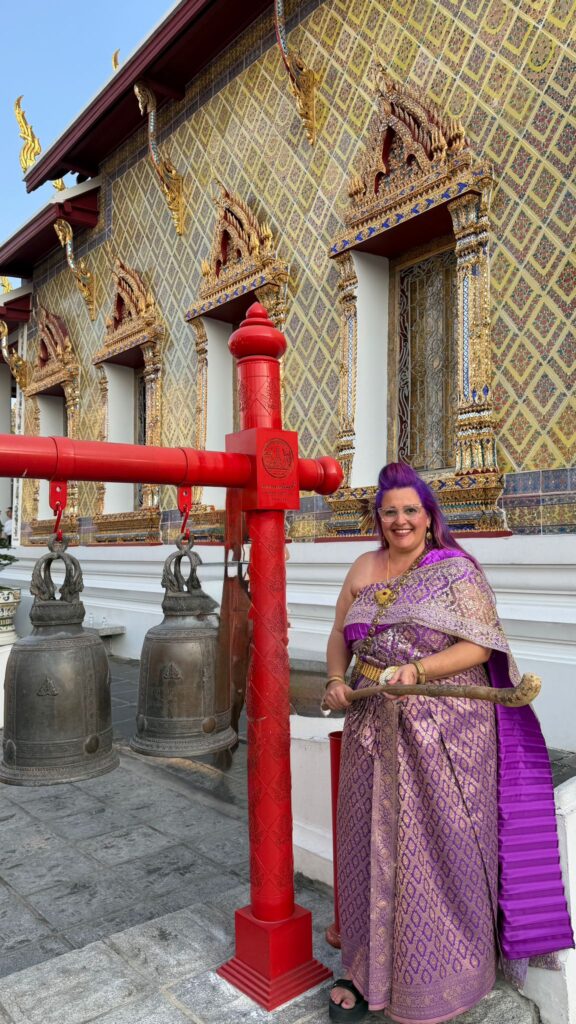 אישה מחייכת בשמלה מסורתית סגולה וזהובה עומדת ליד פעמונים גדולים תלויים ומוט אדום במקדש מעוטר בעושר, עם דוגמאות זהב מורכבות וחלונות מעוטרים.