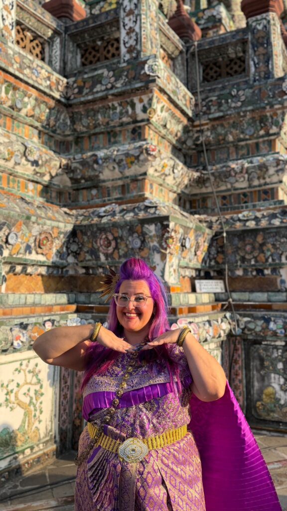 אדם מחייך עם שיער סגול בוהק ומשקפיים מצטלם עם ידיו מתחת לסנטרו, לבוש בבגד מסורתי סגול, מול קיר מקדש מעוטר ומואר בשמש.