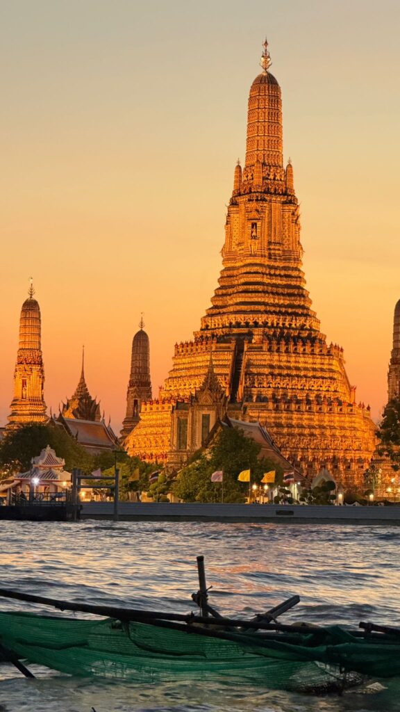 מקדש וואט ארון בבנגקוק, תאילנד, מואר בשקיעה עם שמיים זהובים ברקע. הנהר נראה בקדמת התמונה, עם סירה הנראית בחלקה.