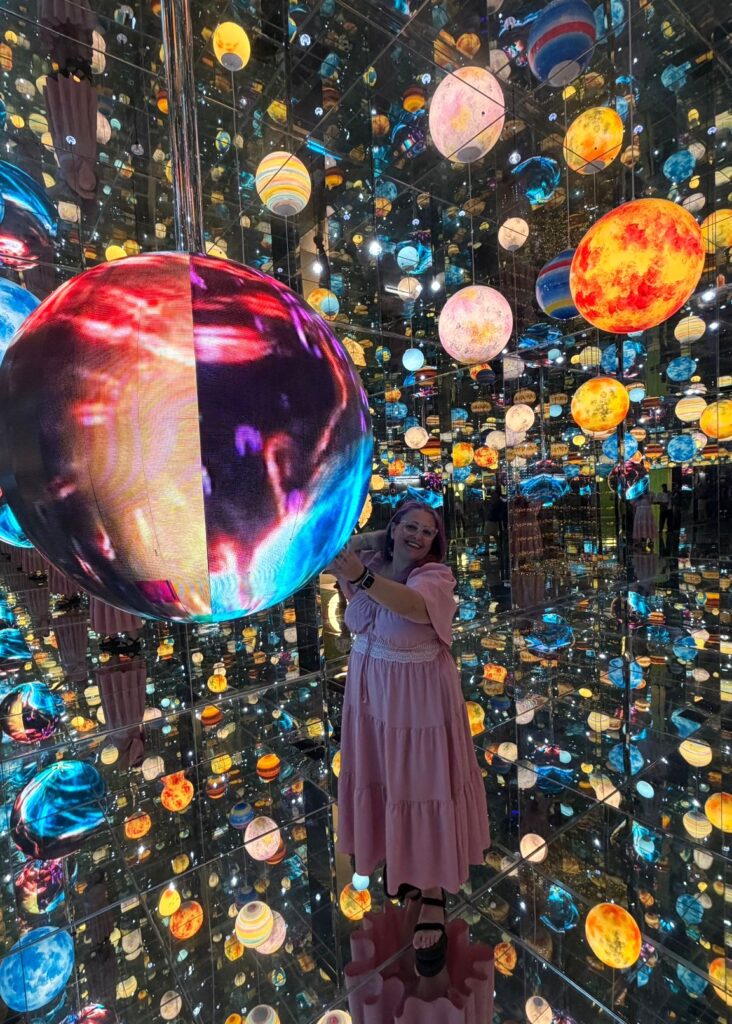 אישה בשמלה ורודה עומדת בחדר מרופד במראות, מלא בכדורים צבעוניים ומוארים הדומים לכוכבי לכת, ויוצרים אפקט של מרחב אינסופי ומרתק. היא מחייכת ומצטלמת עם אחד הכדורים הגדולים.
