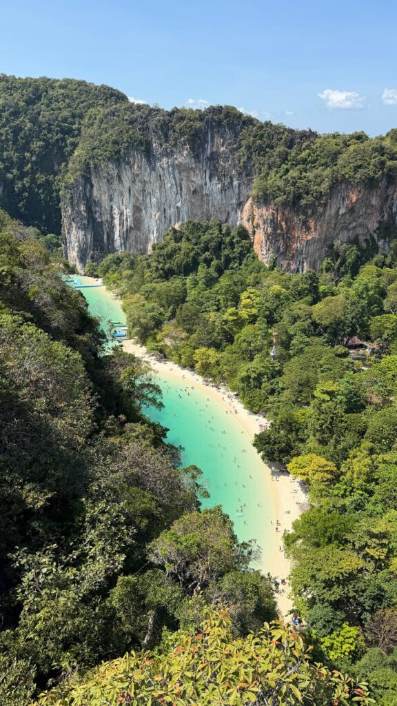 חוף טרופי עם מים בצבע טורקיז, חול לבן ואנשים שוחים, מוקף ביער ירוק צפוף וצוקי גיר גבוהים תחת שמיים כחולים וצלולים.