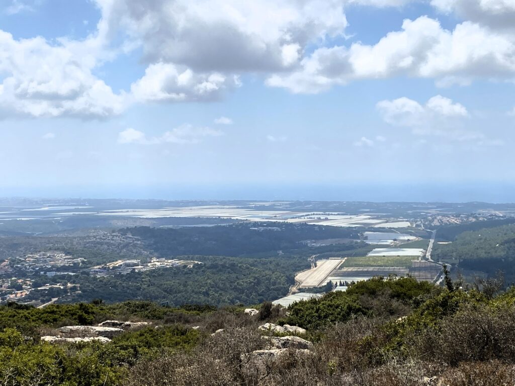 נוף מגבעה המשקיפה על עמק עם שדות ירוקים, מבנים פזורים וכתמי יער תחת שמים מעוננים חלקית, כאשר האופק הרחוק נפגש עם הים.
