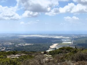 נוף מגבעה המשקיפה על עמק עם שדות ירוקים, מבנים פזורים וכתמי יער תחת שמים מעוננים חלקית, כאשר האופק הרחוק נפגש עם הים.