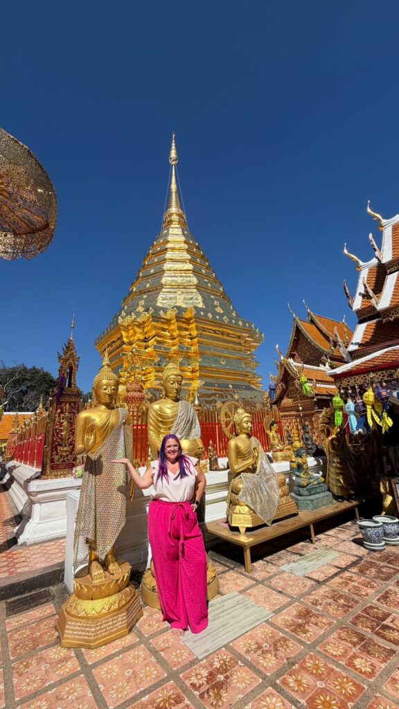 אישה בחצאית ורודה עומדת מחייכת בין פסלי בודהה מוזהבים מול פגודה מוזהבת במקדש, עם שמיים כחולים צלולים מעל ומבני מקדש מעוטרים ברקע.
