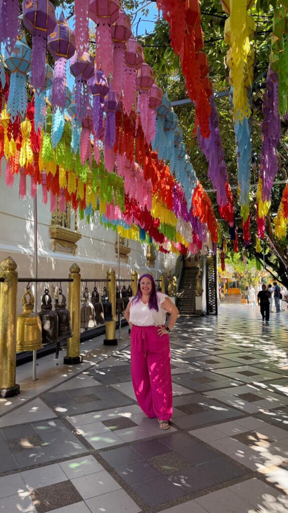 אישה עם שיער סגול ומכנסיים ורודים בוהקים עומדת מתחת לפנסים צבעוניים תלויים מנייר בשביל חיצוני מואר בשמש, כשברקע פסלי בודהה מוזהבים מסודרים בשורה לאורך קיר לבן.