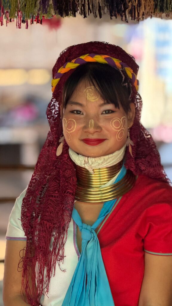 אישה צעירה עם טבעות פליז על צווארה, צבעי פנים מסורתיים וצעיף אדום על ראשה מחייכת למצלמה. היא לובשת בגד אדום-לבן עם צעיף כחול, עונדת עגילים ויש לה ציורים מורכבים על הלחיים.