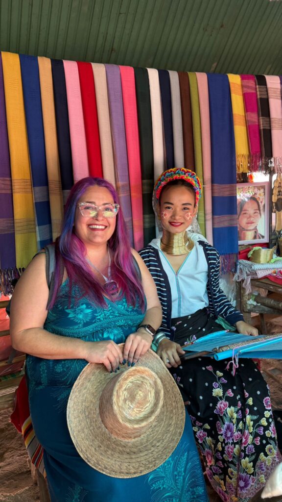 שתי נשים יושבות יחד מול תצוגה של צעיפים צבעוניים ארוגים. לאחת מהן שיער צבעוני, משקפיים, והיא מחזיקה כובע קש, בעוד השנייה לובשת לבוש מסורתי ותכשיטים עם טבעת צוואר מפליז.