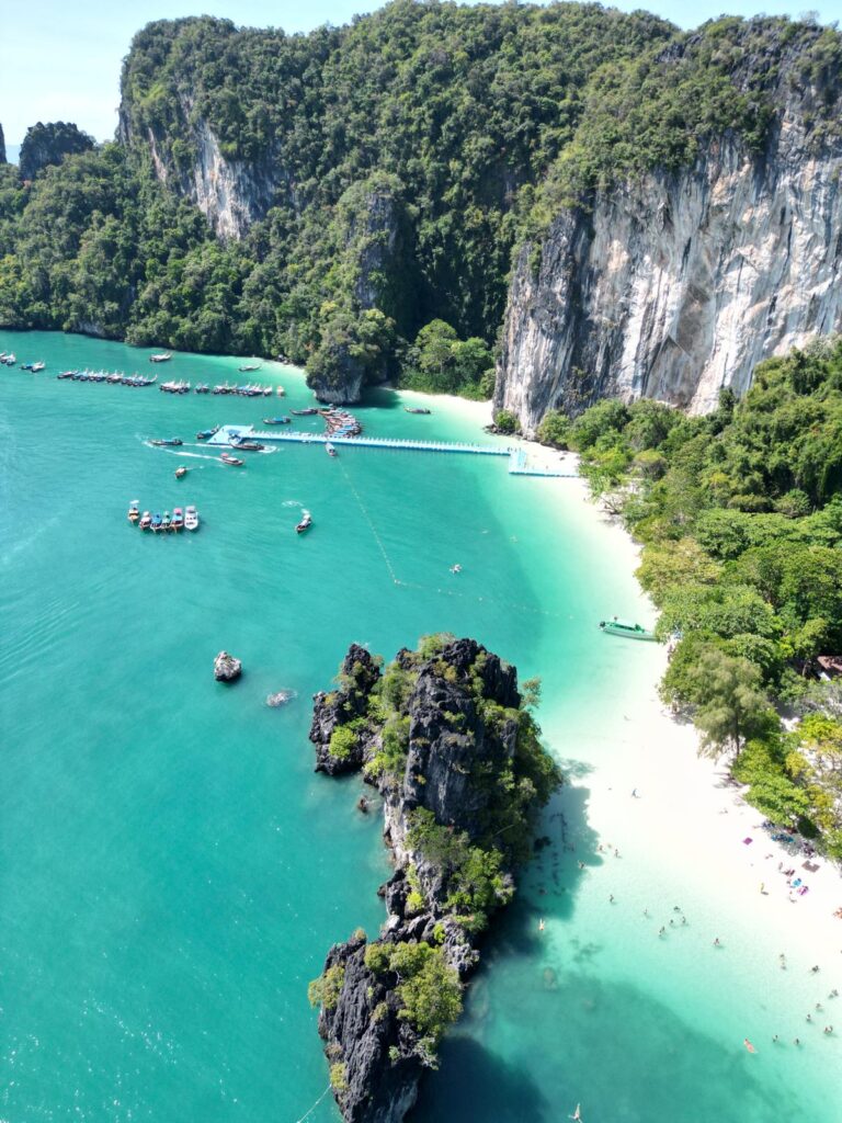 מבט אווירי על חוף טרופי עם מים בצבע טורקיז, חול לבן, צוקים ירוקים ושופעים, סירות עוגנות במזח ואנשים המשתזפים על החוף תחת שמש בהירה.