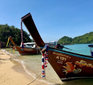 שתי סירות עץ ארוכות זנב, מקושטות בזר פרחים צבעוניים, עוגנות על חוף חולי עם מים כחולים וצלולים. ברקע נראים גבעות ירוקות ושופעות ושמיים כחולים.