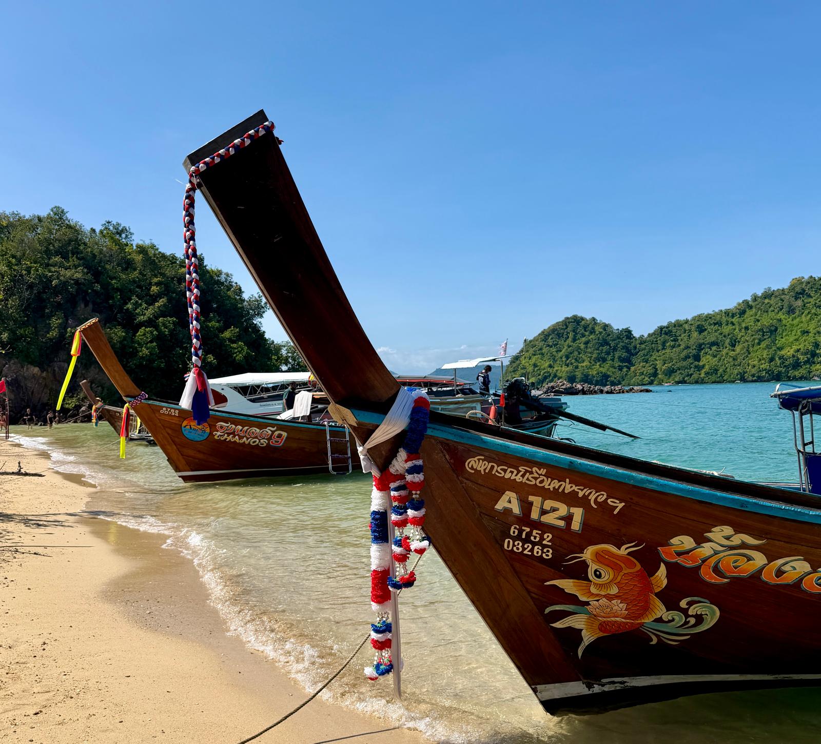 שתי סירות עץ ארוכות זנב, מקושטות בזר פרחים צבעוניים, עוגנות על חוף חולי עם מים כחולים וצלולים. ברקע נראים גבעות ירוקות ושופעות ושמיים כחולים.