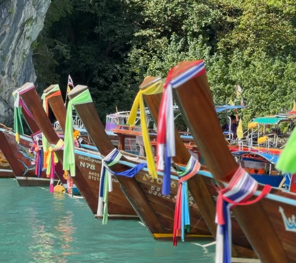 שורה של סירות עץ מסורתיות עם זנב ארוך, מקושטות ברצועות בד צבעוניות, עוגנות לצד מים בצבע טורקיז, על רקע עצים ירוקים ושופעים וצוק סלעי.