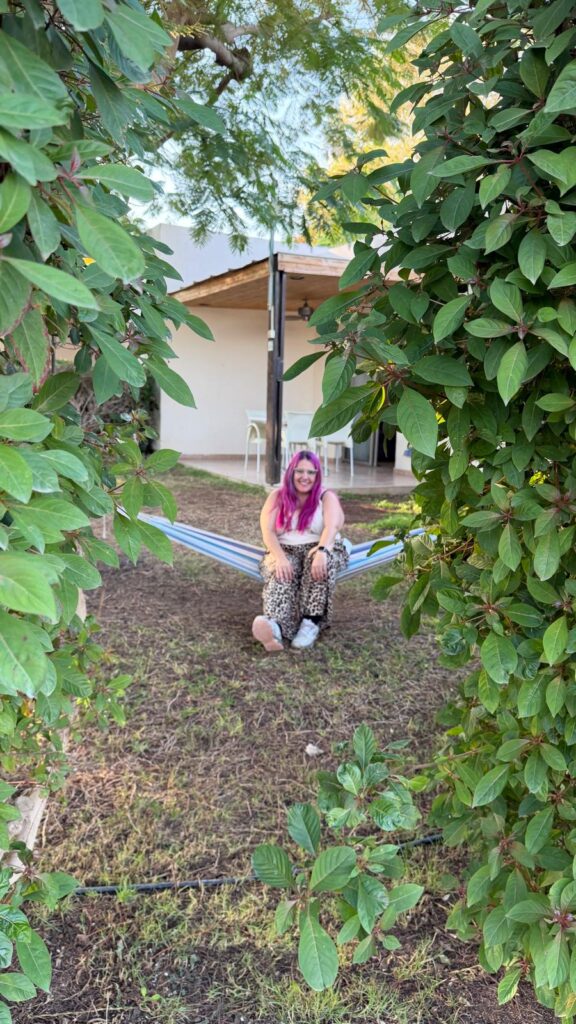 אדם עם שיער ורוד ומכנסיים מעוצבים יושב על ערסל בגינה, מוקף בענפים ירוקים ועלים. ברקע ניתן לראות בית עם פטיו מקורה וכיסאות לבנים.