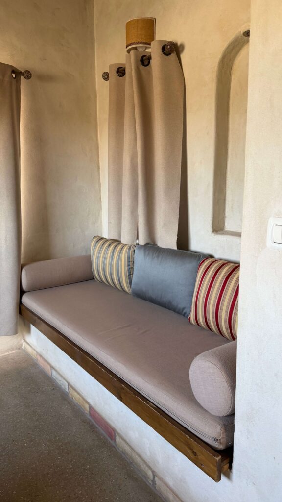 ספסל מובנה ונעים עם ריפוד בצבע בז' ניצב מול קיר כפרי בצבע שמנת, מעוטר בכריות פסים וכריות אחידות ומוקף בווילונות בצבע בז'.