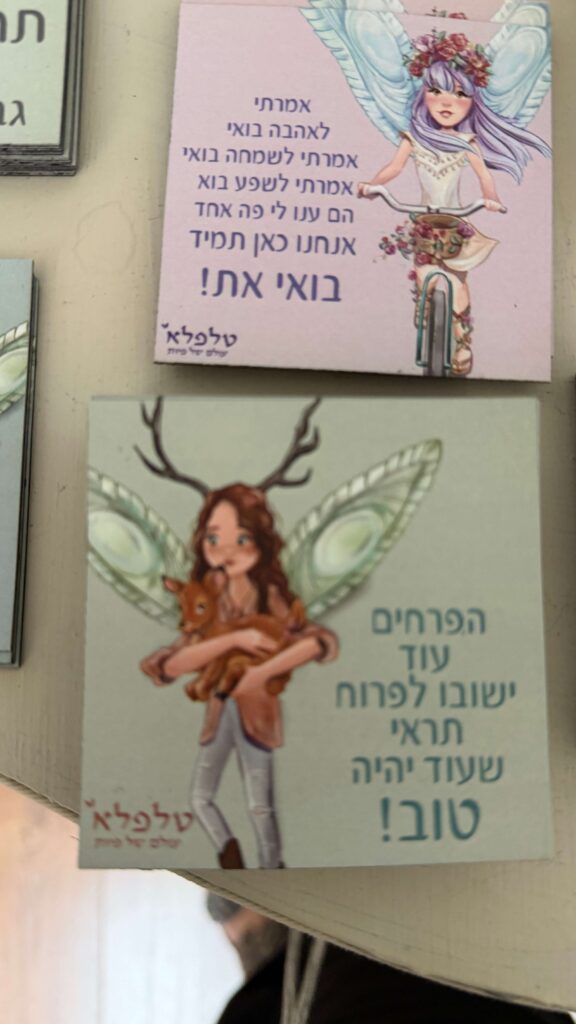 תמונה של שני כרטיסים מאוירים עם טקסט בעברית. הכרטיס העליון מציג ילדה עם שיער סגול רוכבת על אופניים, והכרטיס התחתון מציג פיה עם קרניים המחזיקה גורי צבי. שני הכרטיסים מעוטרים באיורים דקורטיביים ושובבים.