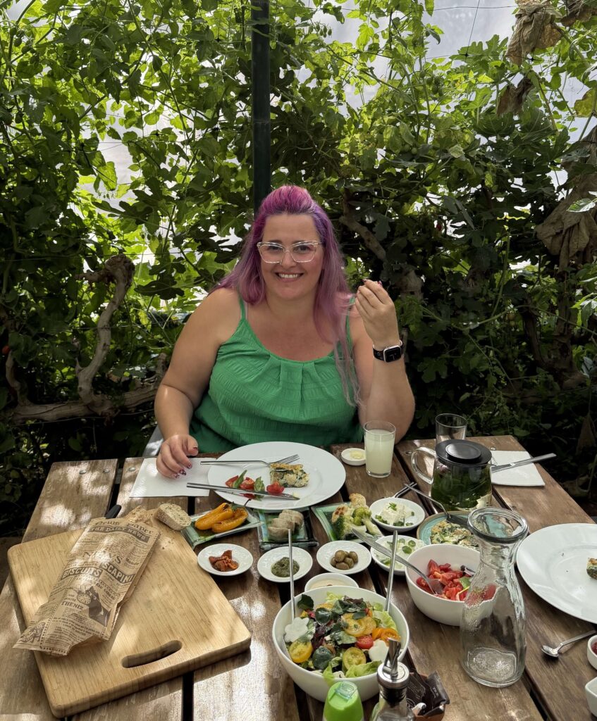 אישה עם שיער ורוד ארוך ומשקפיים, לבושה בחולצה ירוקה, יושבת ליד שולחן עץ בחוץ, מוקפת בצמחייה. היא מחייכת למצלמה כשעל השולחן לפניה צלחות עם מגוון מאכלים ומשקאות.