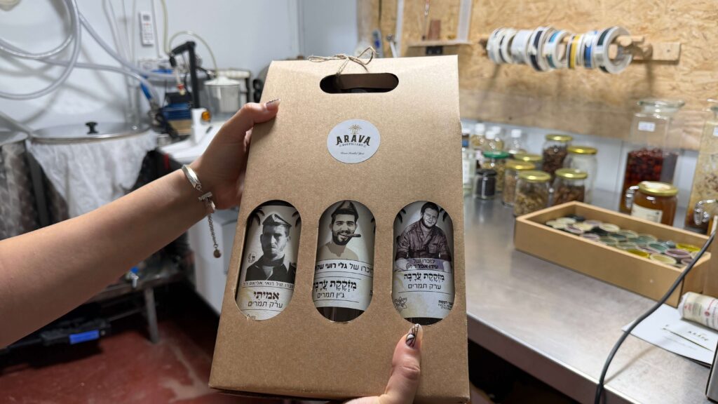 אדם מחזיק בקרטון עם שלוש בקבוקים בתוכו, על כל אחד מהם תווית עם תמונת פניו של גבר. על הקרטון כתוב ARAVA. ברקע נראים צנצנות וציוד על מדפים ודלפקים.