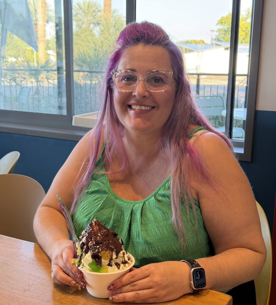 אישה מחייכת עם שיער סגול ארוך ומשקפיים יושבת ליד שולחן בתוך הבית, אוחזת בקינוח עם סירופ שוקולד וקצפת. היא לובשת חולצה ירוקה ושעון חכם, ומאחוריה חלון ונוף חיצוני.