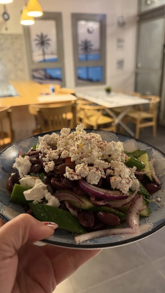 יד אוחזת בצלחת סלט יווני עם גבינת פטה, זיתים, בצל סגול ופלפלים ירוקים. ברקע נראים שולחנות, כיסאות ותמונות ממוסגרות על הקיר במסעדה נעימה ומוארת באור רך.