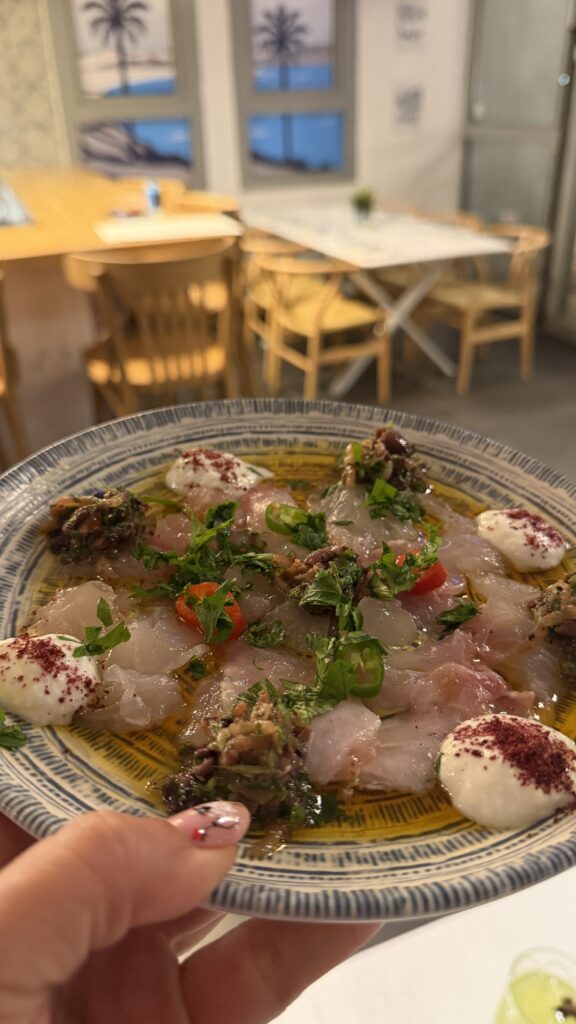 יד אוחזת בצלחת עם פרוסות דקות של דג נא, מעוטרות בכמות נדיבה של רוטב שמנת, עשבי תיבול, שמן זית ותבלינים. ברקע נראית מסעדה נעימה עם כיסאות עץ, שולחנות ועיצוב בסגנון חוף הים.