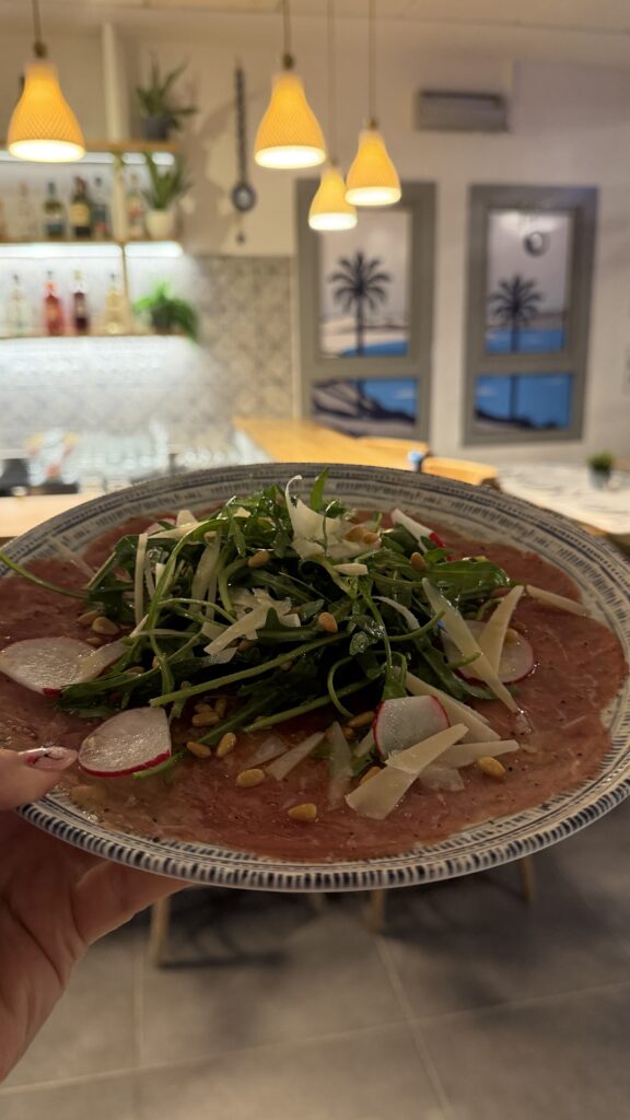 יד אוחזת בצלחת קרפצ'יו בקר עם רוקט, פרמזן מגורר, פרוסות צנון וצנוברים במסעדה נעימה ומוארת באור חם, עם עיצוב מודרני ומדבקות עצי דקל על החלונות.