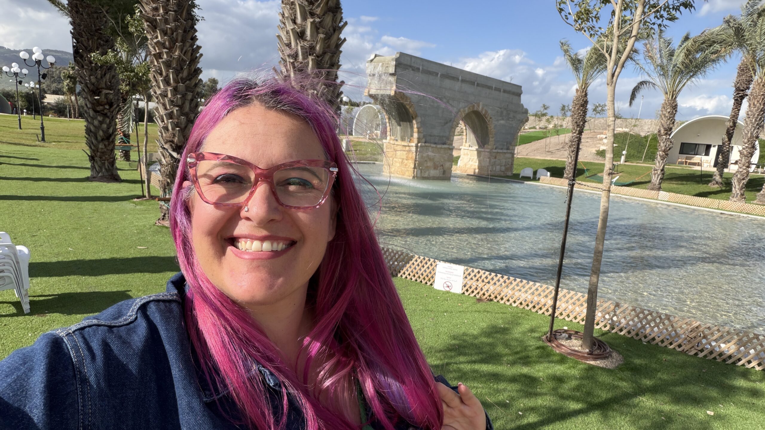 אישה עם שיער ורוד ארוך ומשקפיים מחייכת מול בריכה עם גשר אבן ועצי דקל ביום שטוף שמש.