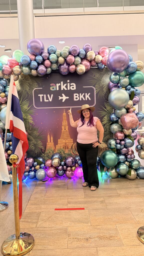 אישה עם כובע מצטלמת ומחייכת מול קשת בלונים דקורטיבית ושלט שעליו כתוב arkia TLV → BKK, עם איור של אתרי תיירות בבנגקוק מאחוריה. דגל תאילנד ניצב משמאל.