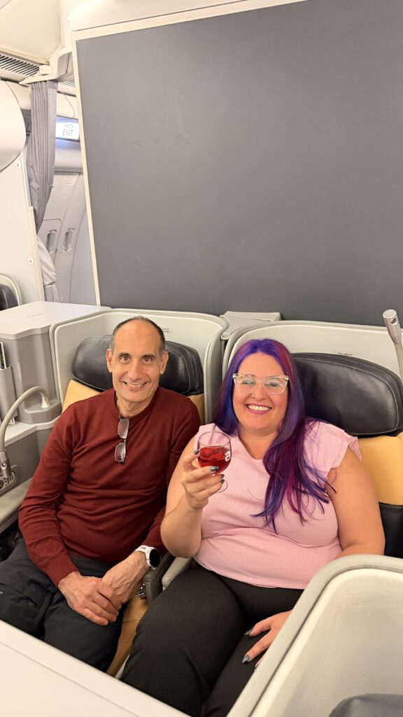 גבר ואישה יושבים במושבים במחלקת העסקים של מטוס; האישה מחייכת ומחזיקה משקה, והגבר מחייך וידיו מונחות על ברכיו.