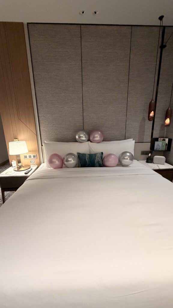 מיטה מסודרת עם סדין לבן וכריות מעוטרת בחמישה בלונים – שלושה ורודים ושניים כסופים – על רקע ראש מיטה מרופד בחדר מלון מודרני ומואר באור רך.