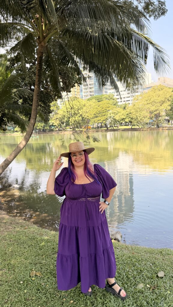 אישה בשמלה סגולה וכובע קש עומדת מחייכת ליד אגם, כשעצים ובניינים גבוהים משתקפים במים מאחוריה ביום שטוף שמש.