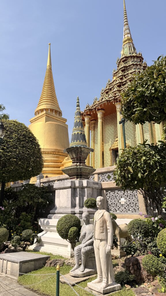 פגודות זהובות ומבני מקדש מעוטרים תחת שמיים כחולים בגן נוי שופע, עם פסלי שיש לבנים של אנשים בחזית, בוואט פרה קאו, בנגקוק, תאילנד.