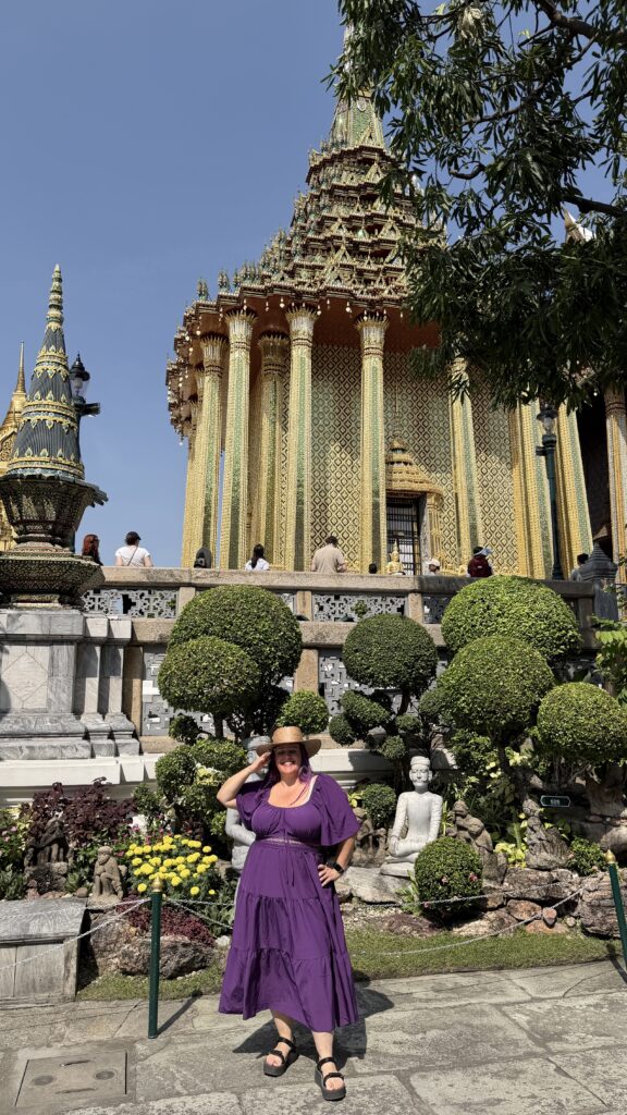 אישה בשמלה סגולה וכובע שמש מצטלמת מול מקדש זהוב ומקושט, מוקפת בגנים מטופחים, פסלים ומבקרים אחרים תחת שמיים כחולים וצלולים.