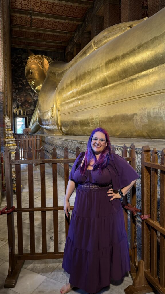 אישה מחייכת עם שיער סגול ושמלה סגולה עומדת יחפה מול פסל בודהה השוכב הענק מזהב בוואט פו בבנגקוק, תאילנד. מעקה עץ מפריד בינה לבין הפסל.