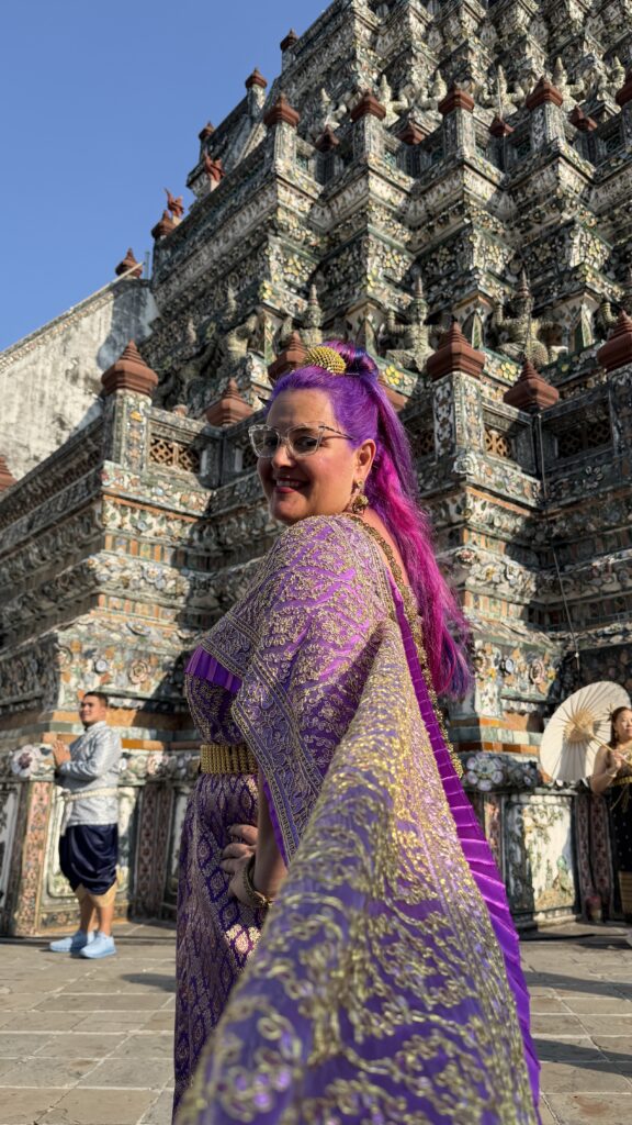 אישה עם שיער סגול בוהק ומשקפי שמש, לבושה בבגד מסורתי מעוטר בסגול וזהב, מצטלמת מול מקדש עתיק ומפורט עם גילופים מורכבים. היא מחייכת ומחזיקה את ידו של מישהו.