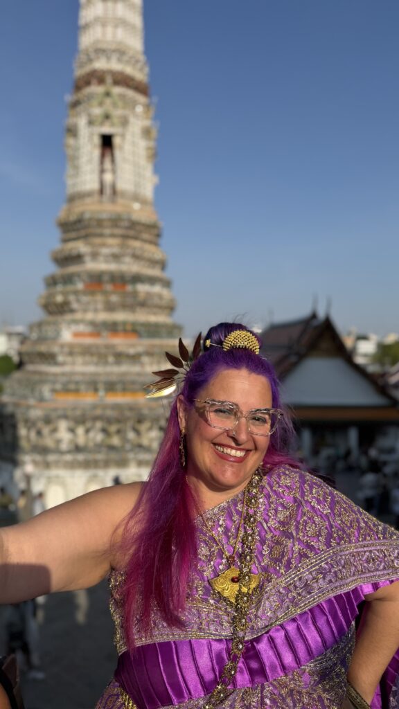 אישה עם שיער סגול בוהק ומשקפיים, מחייכת ולבושה בבגד מסורתי סגול וזהוב, מצטלמת מול מבנה מקדש גבוה ומקושט תחת שמיים כחולים וצלולים.