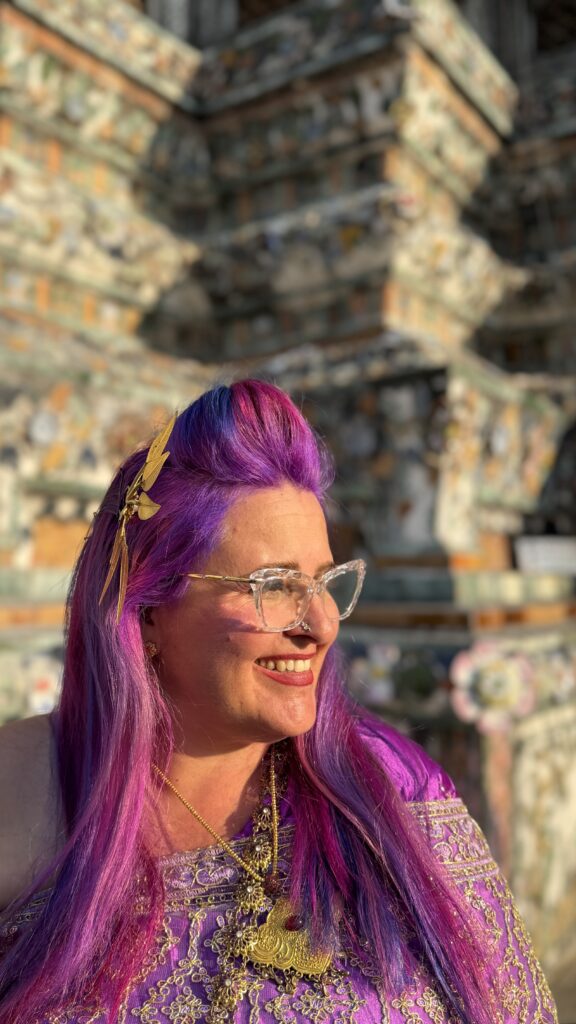 אישה עם שיער ארוך סגול וורוד, עונדת תכשיטי זהב ומשקפיים שקופים, מחייכת בעודה עומדת בחוץ מול מבנה אבן מפורט ומואר בשמש. היא לבושה בבגד סגול עם נגיעות זהב.