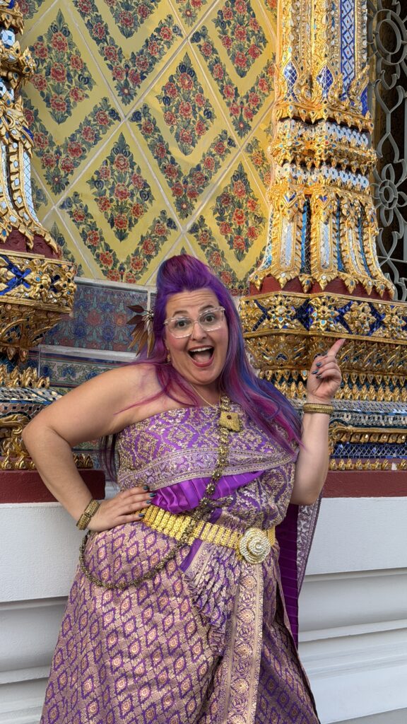 אישה מחייכת עם שיער ורוד וסגול, לבושה בתלבושת מסורתית מעוטרת בסגול וזהב, מצביעה בהתרגשות על קיר זהוב מעוטר בדוגמאות צבעוניות.