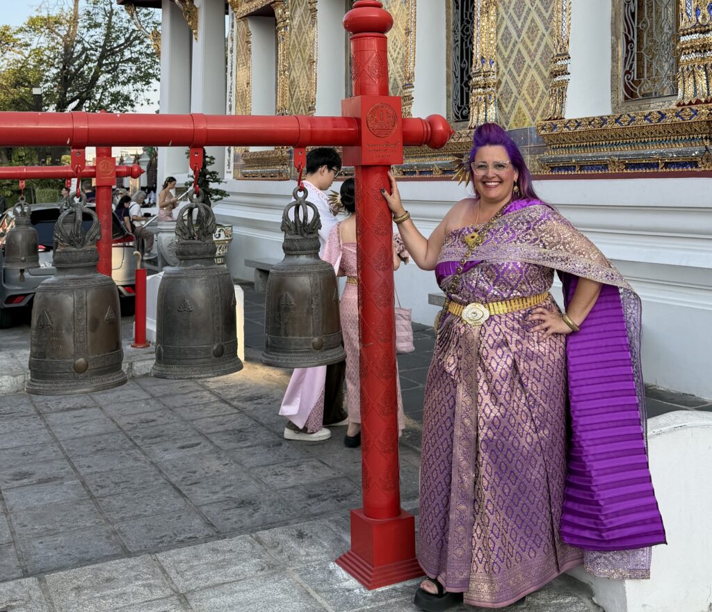 אישה מחייכת לבושה בשמלה תאילנדית מסורתית בצבעי סגול וזהב עומדת ליד פעמונים טקסיים גדולים על מסגרות אדומות מחוץ למקדש מעוטר, כשברקע אנשים אחרים לבושים בבגדים דומים ופרטי מקדש מעוטרים.